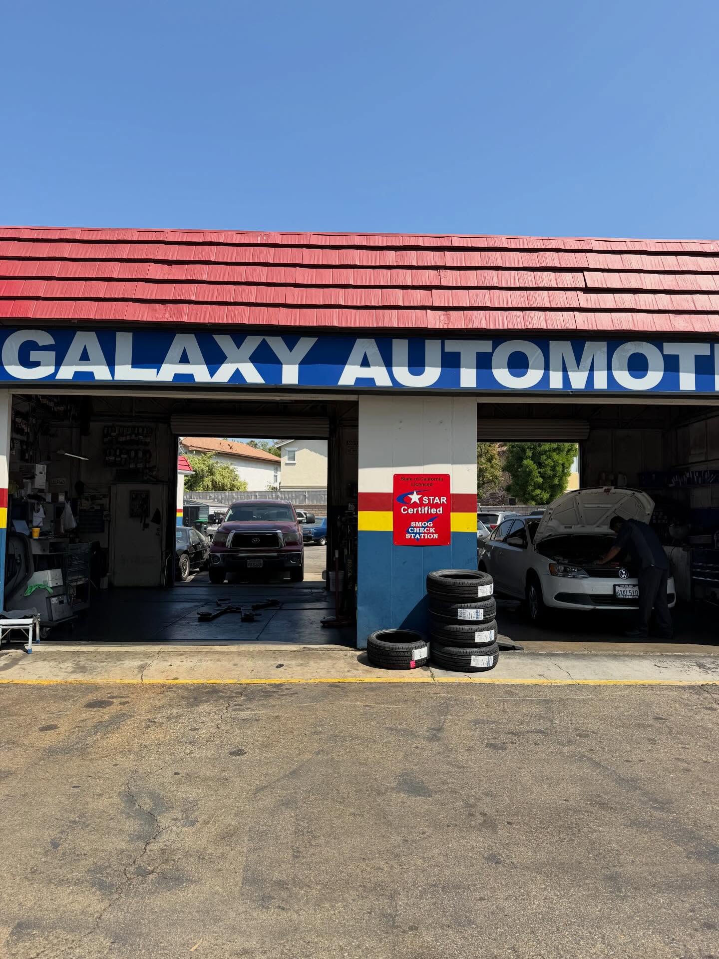 The Galaxy Automotive frontage showing the State of California licensed STAR certified smog check station sign, stacked tires and a technician working in the bay